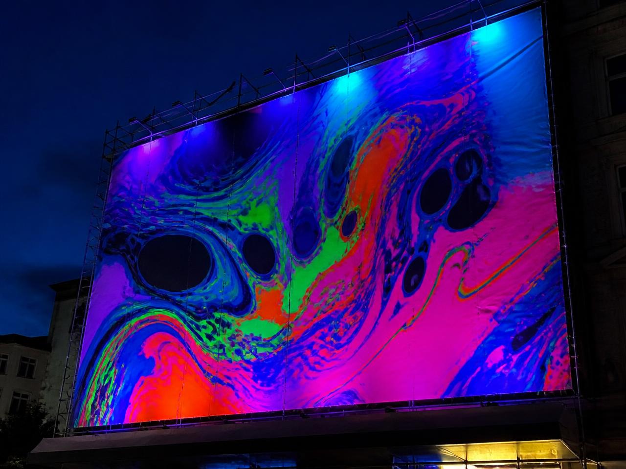 Stort stilladsbanner med fluorescerende print, der gløder under UV-lys om aftenen